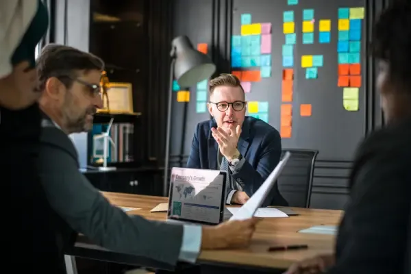 Consultants and clients in a working session around a conference table with growth analytics on a laptop and a sticky-note planning wall.