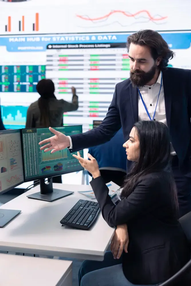 Two consultants collaborating at monitors showing charts and KPIs in a statistics hub with large wall-mounted data displays.