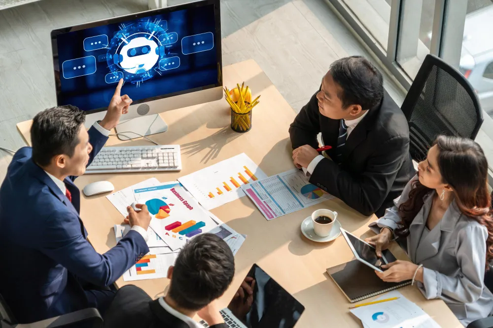 Team of professionals around a desk discussing reports with an AI chatbot visual on a large monitor.