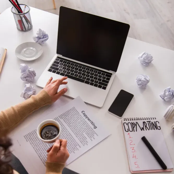 Top-down copywriter desk with a laptop, coffee, a Copywriting brief, notebook checklist and crumpled drafts.
