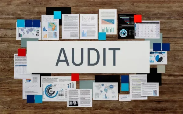 Desk covered with reports, charts and sticky notes around a central banner reading AUDIT.