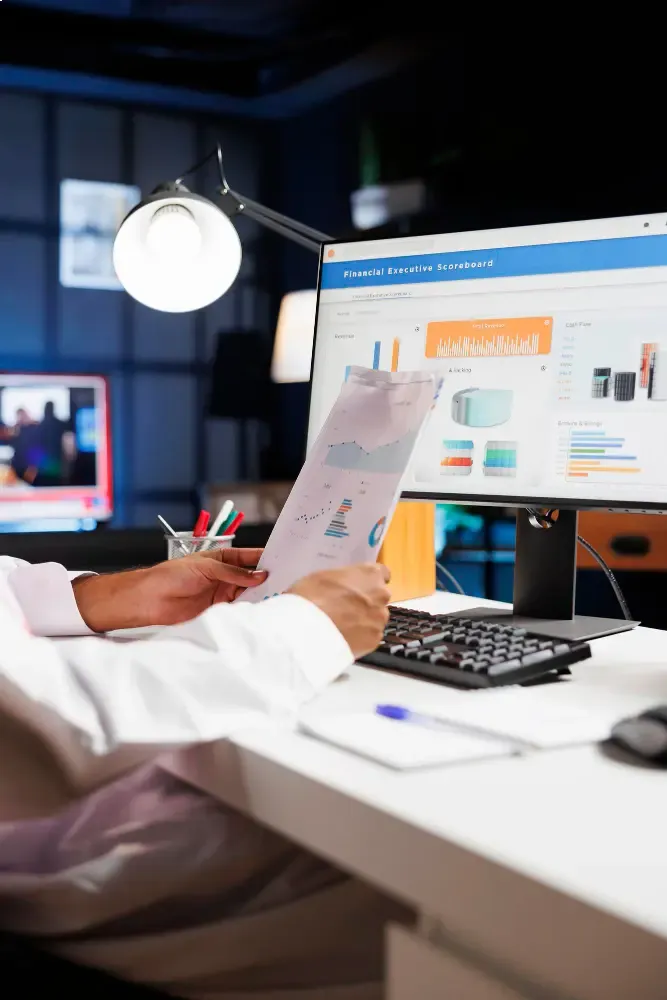 Analyst comparing printed charts with performance dashboards on a large monitor.