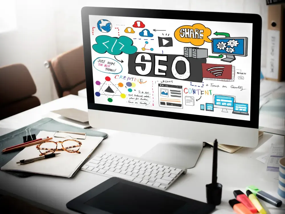 Bright office desk with a monitor showing a hand-drawn SEO infographic—creative content, layout, share, quality themes—plus graphics tablet, notebook with glasses, and markers.