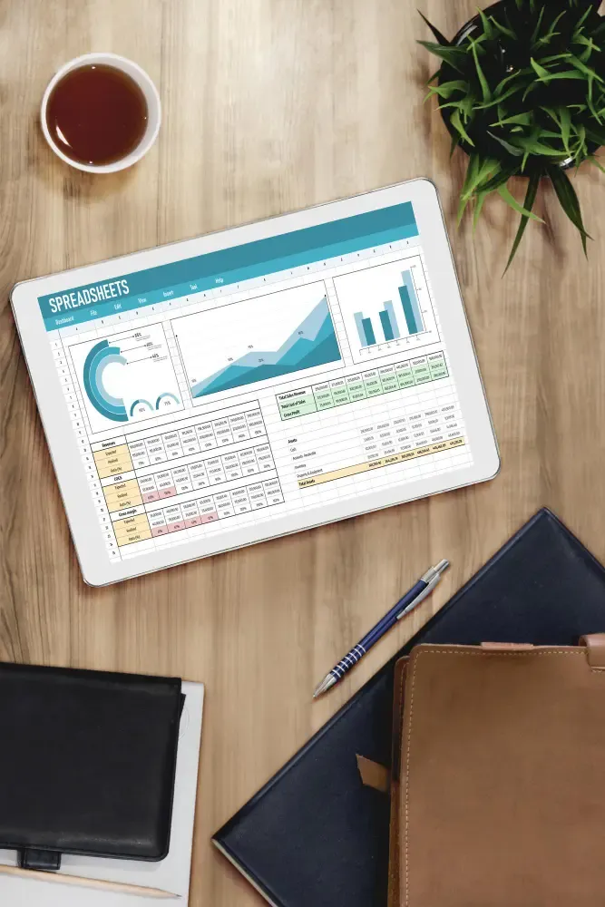 Wooden desk with a tablet showing spreadsheet dashboards—charts and data tables—with coffee, a plant, pen, and notebooks nearby.