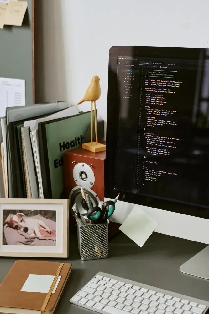 Desk workspace with a monitor showing code in an editor, leather notebook, framed photo, pencil cup, and folders—hands-on refinement of digital content.