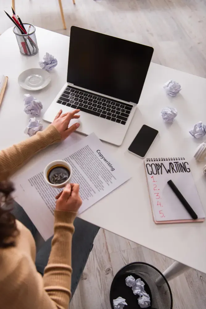 Desk with laptop, printed copywriting draft, notebook labelled COPYWRITING with numbered notes, coffee cup, phone, and crumpled paper drafts.