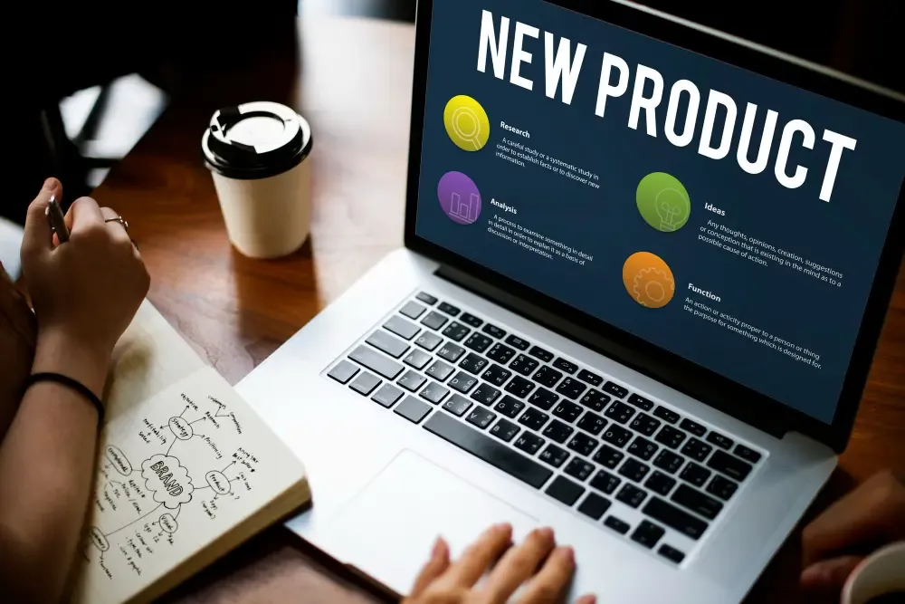 Person at a wooden desk with a laptop showing a New Product infographic—research, analysis, ideas, function—and an open notebook with hand-drawn brand strategy notes.