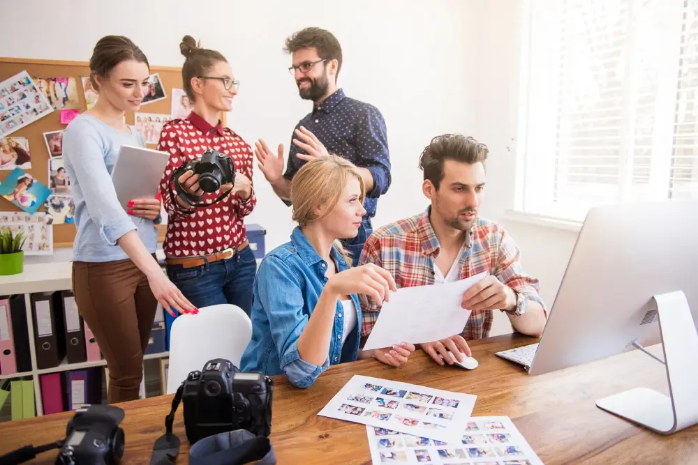 Creative team collaborating around a desk with an iMac, cameras, photo contact sheets, and a corkboard of visuals in a bright studio office.