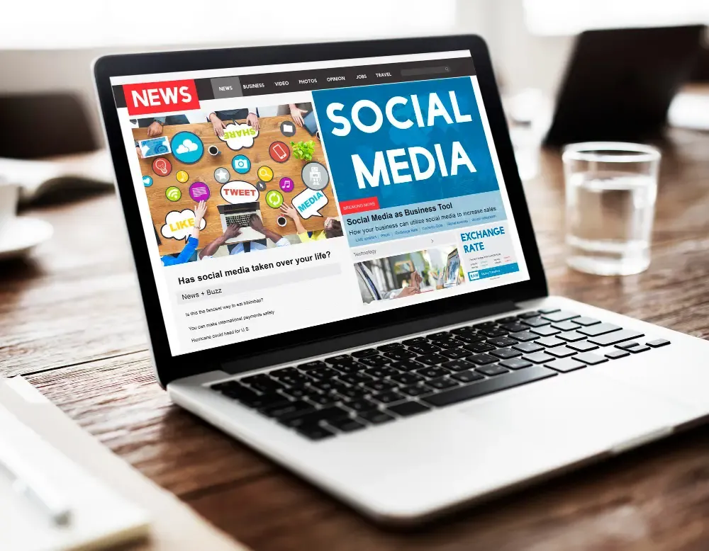 Laptop on a wooden desk showing a news-style webpage with a Social Media banner, articles on social media for business, and illustrated engagement icons.